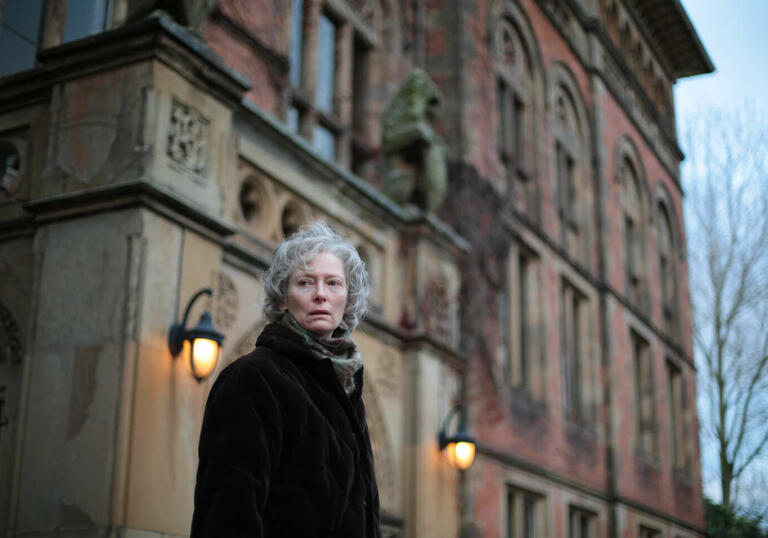 On a winter's day, a woman with grey hair walks away from a large, stately building.