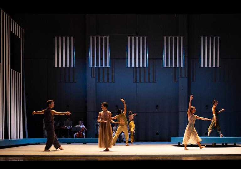 A company of dancers perform on stage in muted coloured clothing.