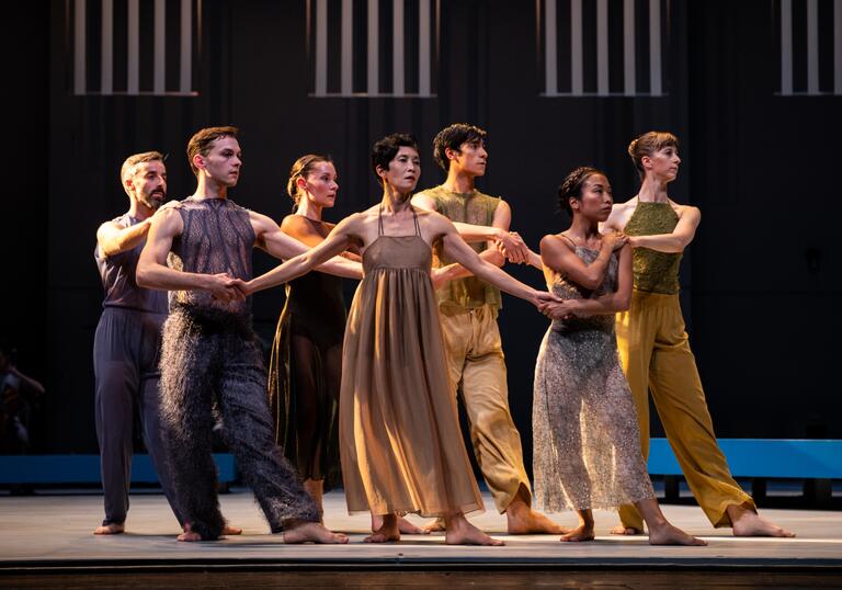 Seven dancers stand arm-in-arm looking off-stage. They are wearing muted colours.