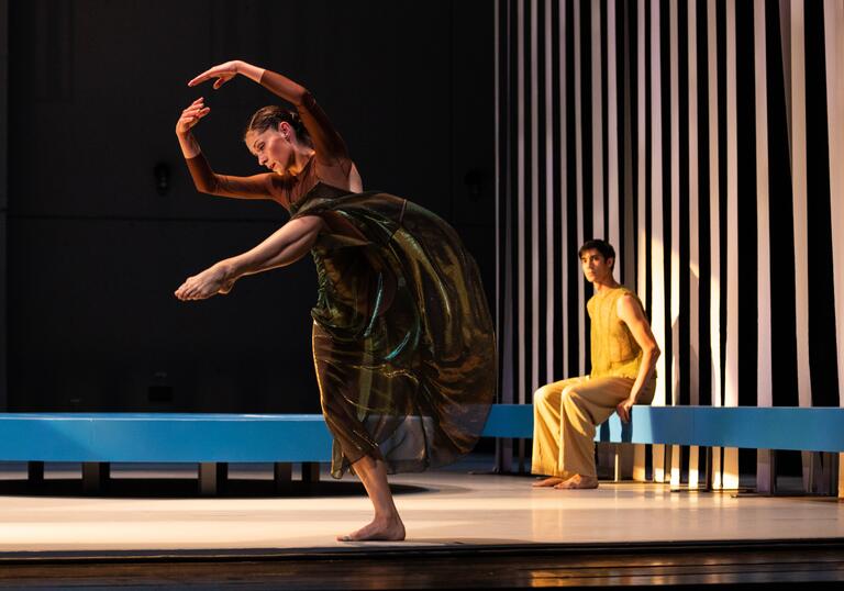 A dancer performs with their arms bowed above their heads and one leg raised in the air. Behind them a person dressed all in yellow sits on a blue bench watching.