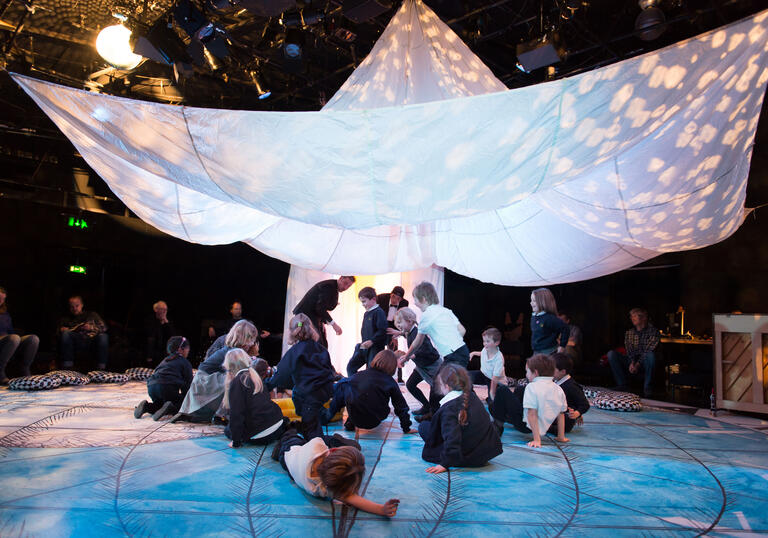 A group of families enjoying a performance of Told by an Idiot's Get Happy. Children gather under a big, white parachute. 