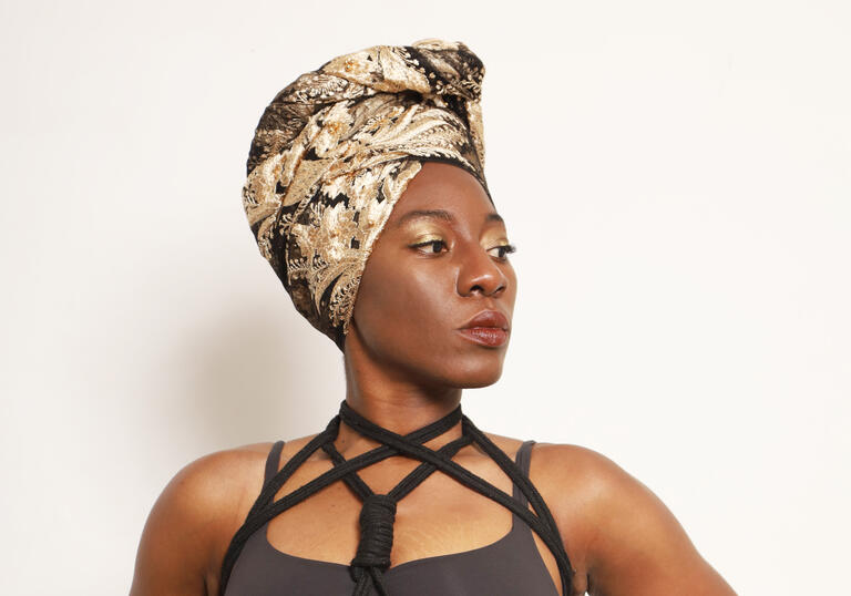 A person wearing a grey bodysuit and a cream, brown and black headscarf stands against a white wall and looks off camera to their left. There are black ropes tied around their chest.