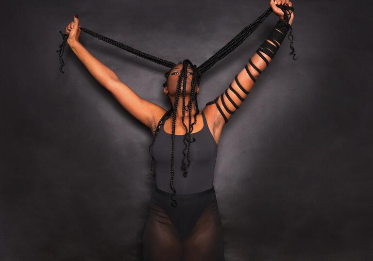 A performer wearing a grey bodysuit stands against a grey background. They hold onto black ropes.