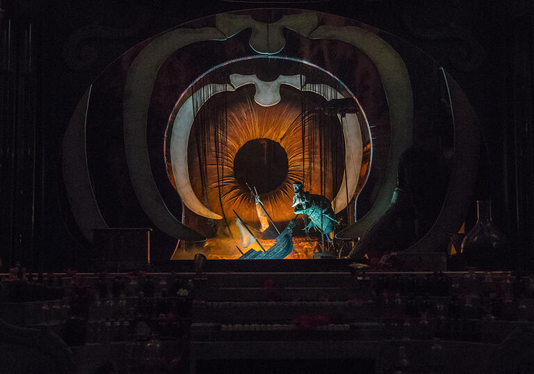 Two puppets, one a creature and one a ship, appear to be falling into a stage. The stage looks like the inside of a circus tent, dark red and yellow in colour.