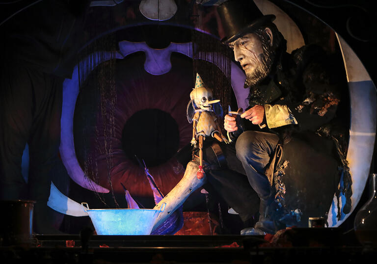 A large puppet person with a beard, wearing a long black coat and a top hat, sits down on stage with a small wooden Pinocchio puppet. 