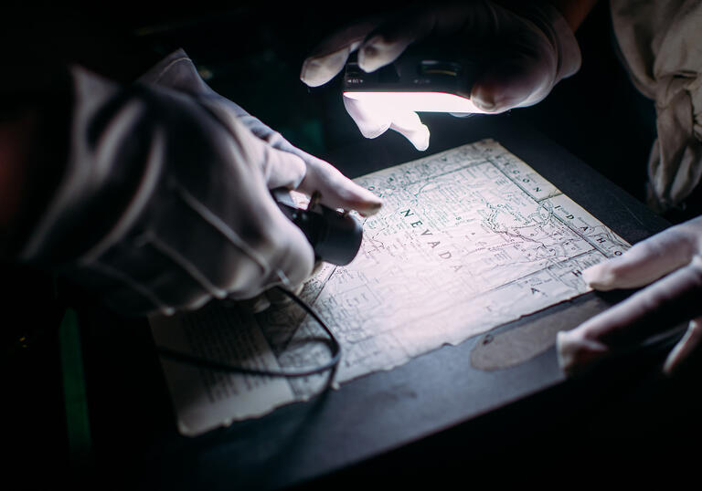 Two pairs of hands wearing white gloves move a camera over a map.