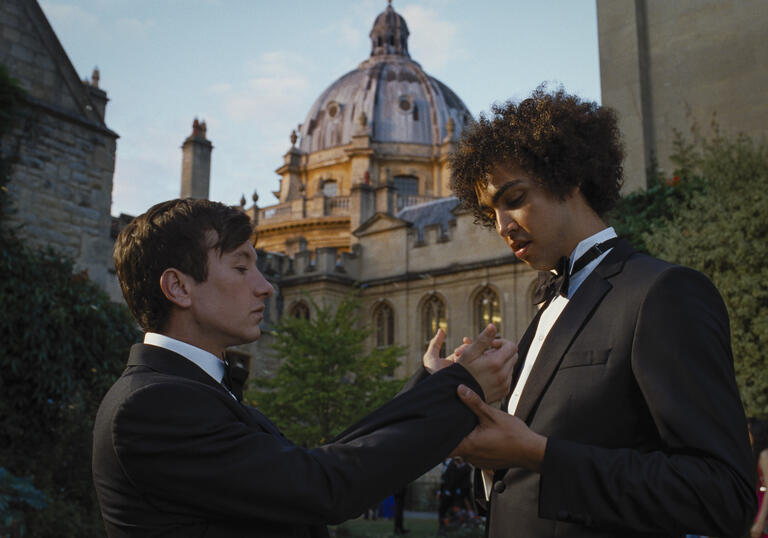A young boy fixes the cuff link of another boy, both in black tie, in front of a grandiose building. 