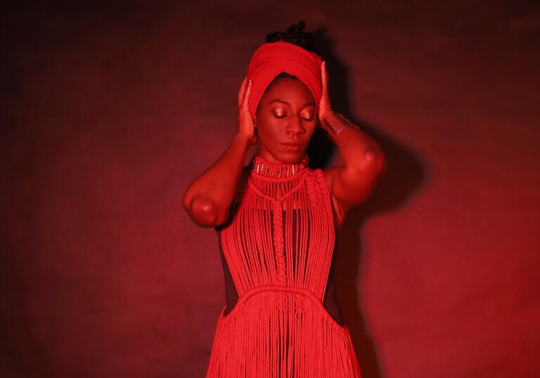 A person wearing a cream dress and matching head scarf closes their eyes and holds their hands on their head as they stand against a red wall.