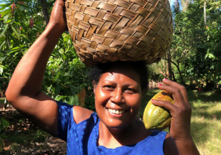 Lucy, who owns a chocolate farm, wears a blue top and carries a basket on her head. She smiles at the camera.