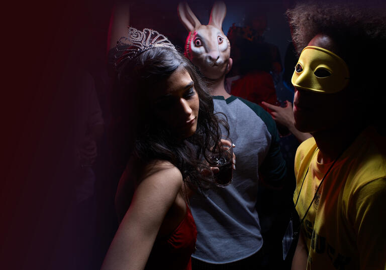 Party scene with person on left wearing a tiara and holding drink, person in middle with rabbit mask on face, person on right with yellow mask over eyes