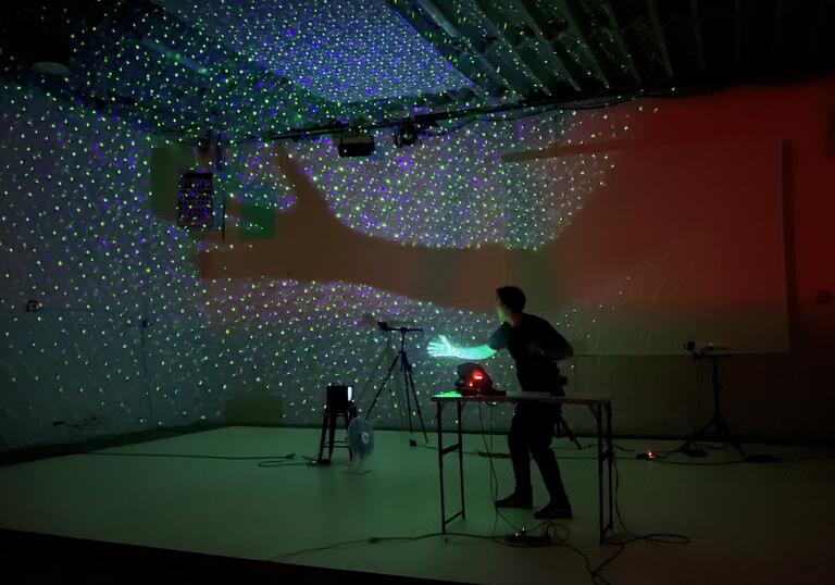 A person stands in a room with a lot of technical equipment around them. Their arm is projected onto a large screen with lots of light.