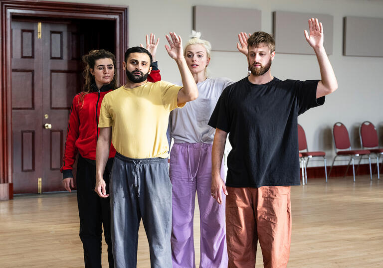 Four people stand in a group with one hand raised in the air as if ready to wave. They have concerned expressions on their faces.