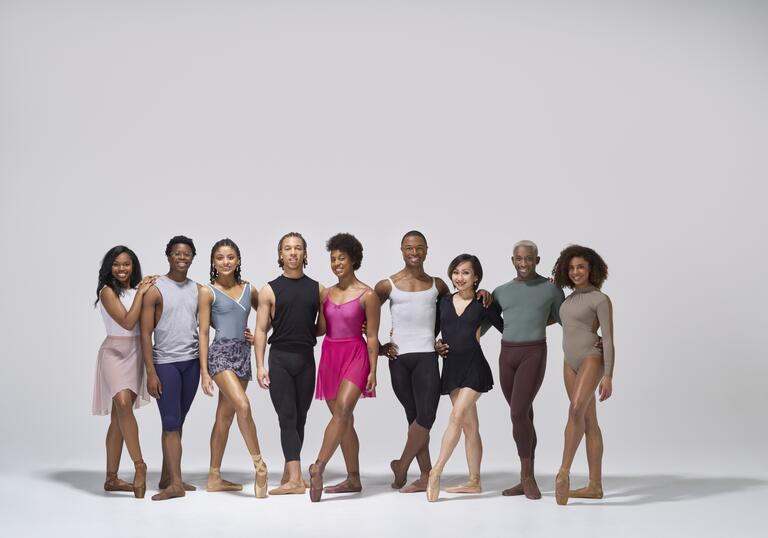 Members of the Ballet Black company pose in a line, smiling. They are standing against a grey background.