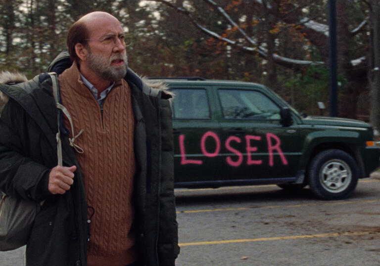Nicholas Cage walks through a car park in front of a car that has LOSER spray painted on it in red.