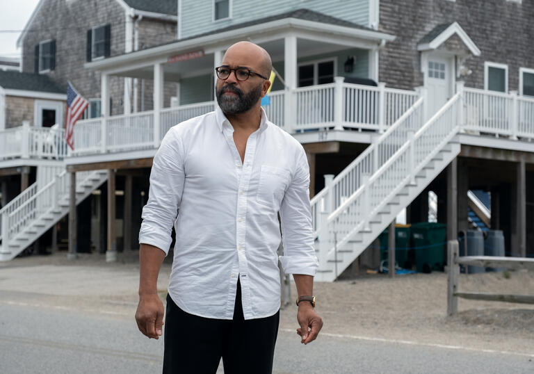 A man in a white shirt and glasses looks out over a parking lot, in front of white buildings. 