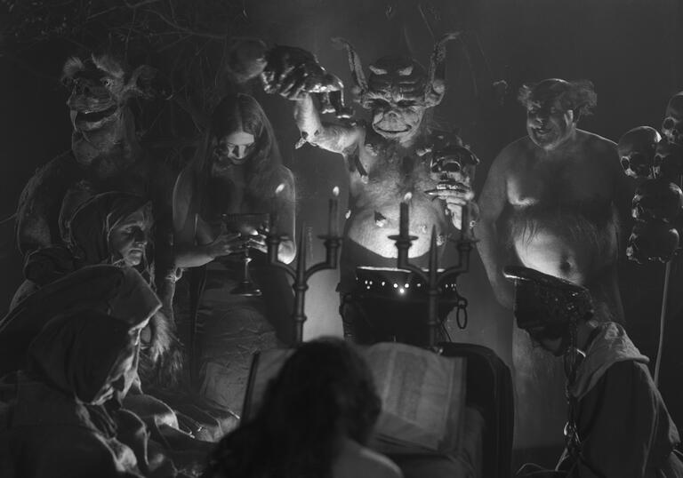 A satanic group gather in dark candlelight.