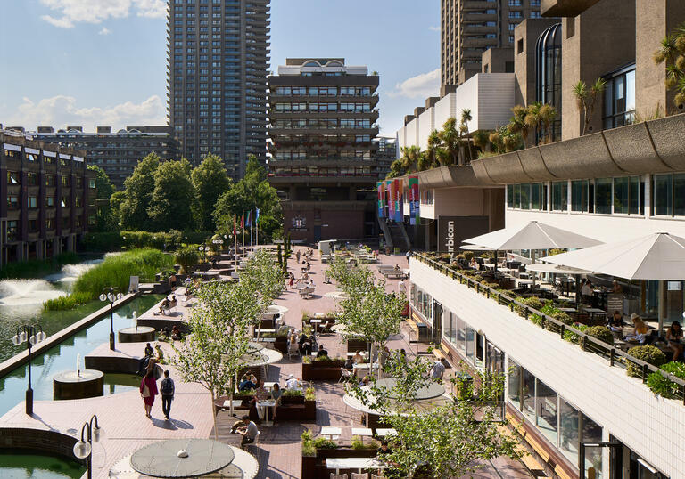 A view in sunshine of Barbican's lakeside terrace, with the lake to the left and trees, tables and people alongside it