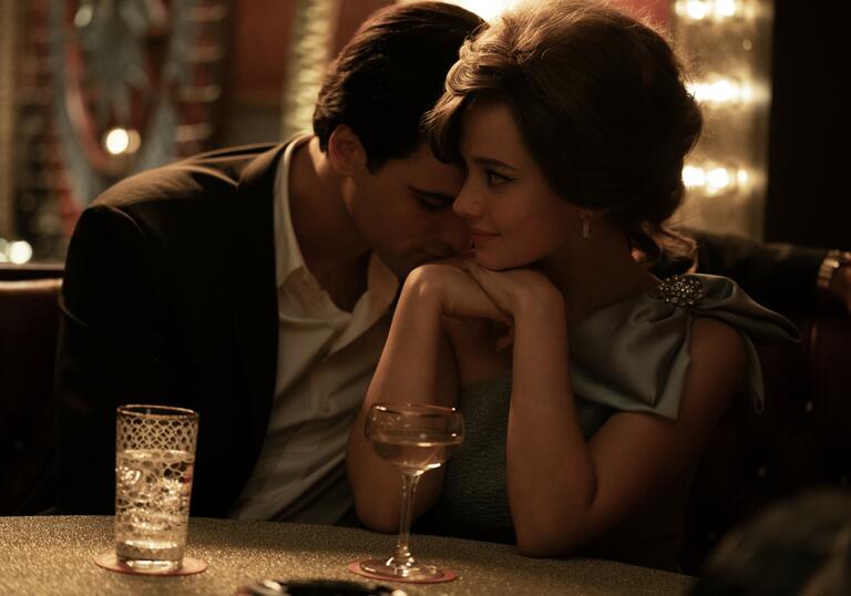 A man rests his head on a young woman's neck at a table in a dark bar.