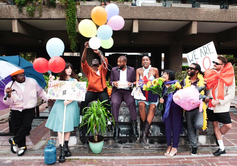 The PappyShow company sit on a wall at the Barbican Centre with signs, balloons, flowers and other objects celebrating the fact they are 10.