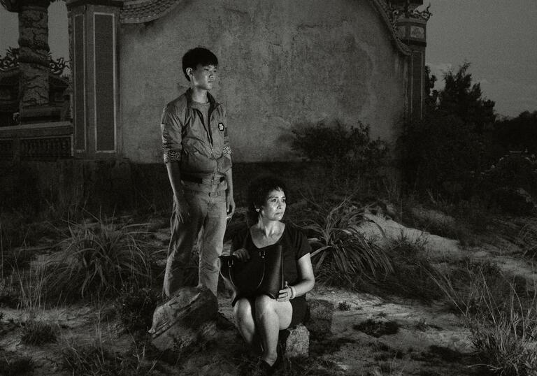 A man and a woman look solemnly forward from a landscape.