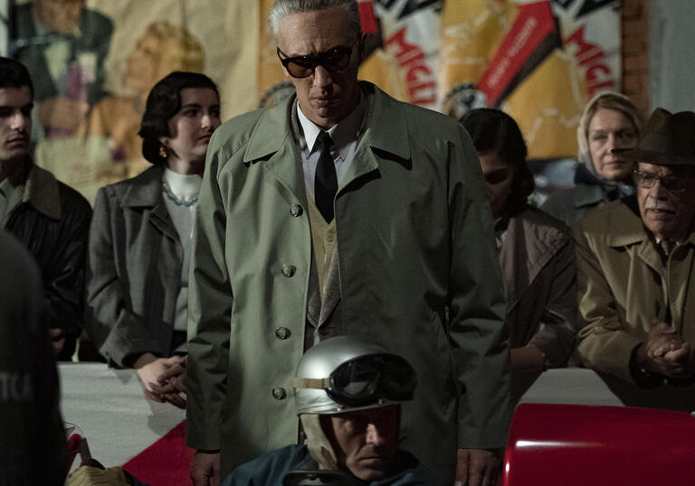 Enzo Ferrari wears glasses on a busy street, in front of a race car.