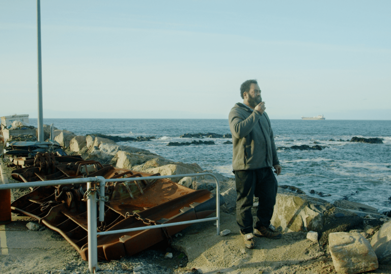 A man stands on a pier smoking a cigarette next to an old boat.