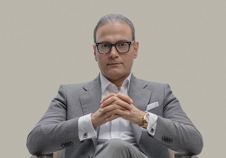 Alireza Ghorbani sits on a wooden chair facing the camera, legs crossed and wearing a grey suit