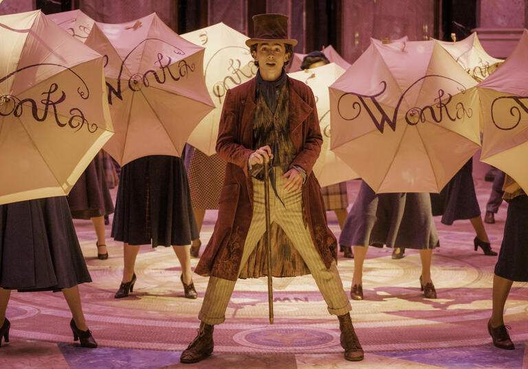 Willy Wonka, wearing a top hat and cane, stands on stage surrounded by umbrellas with 'WONKA' written on them.