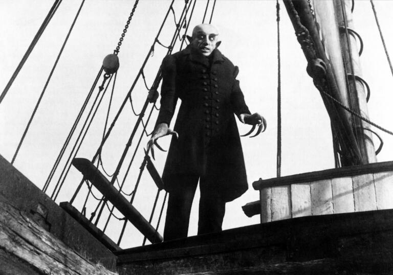 Still from 1922 silent film Nosferatu: A Symphony of Horror, showing Nosferatu with his long, claw-like fingers and pointed ears, standing on a ship wearing a long black coat