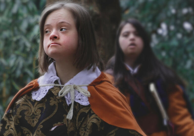 Two young girls in period dress stand stoically in a forest. 