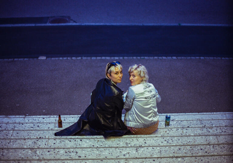 Two people sit on the curb in the dark, looking over their shoulders straight at the camera.
