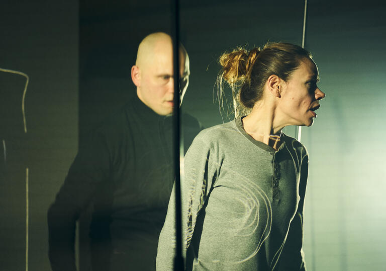 Two performers on stage: one creeps up behind the other menacingly.