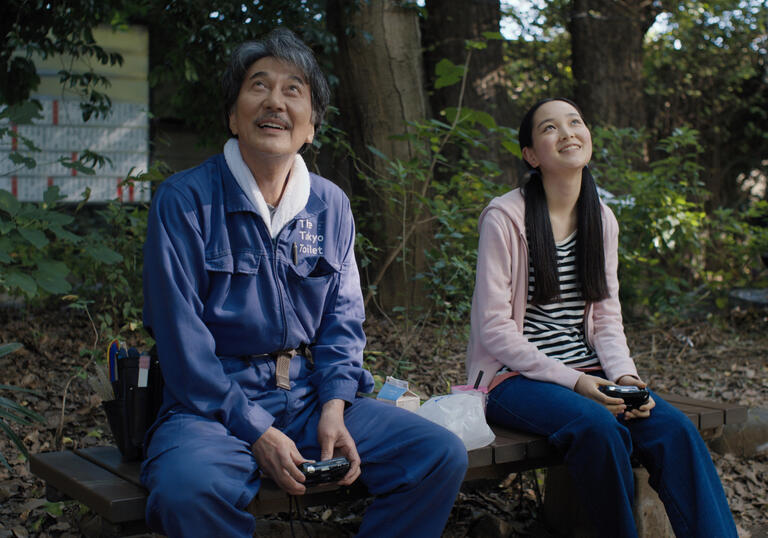 A man and a girl sit on a bench in a park, looking up to the sky and smiling. 