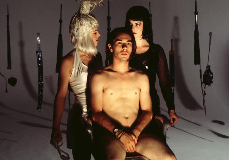 A man sits naked in a chair of a dark room flanked by two women. There are weapons on the wall behind them.