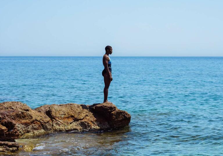 A woman in a bathing suit stands on a rock, looking out across the blue ocean in front of her.