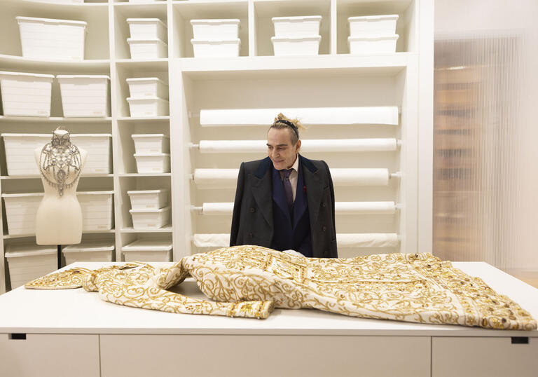 Fashion Designer John Galliano looks down at a beautiful cream gown that lies on a table in a cream room.