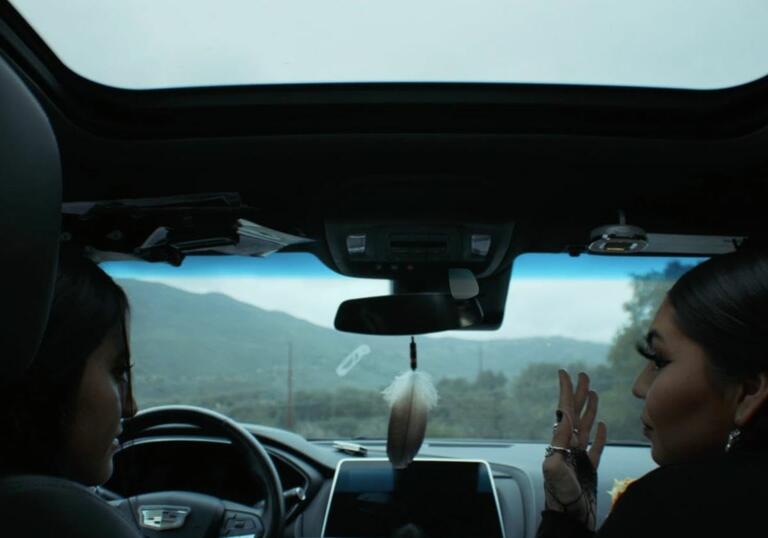 Two women sitting in the front of a car talk to each other, in front of a moody landscape.