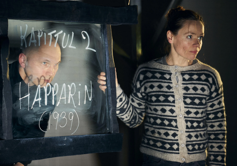 Two people perform in a theatre with a glass screen between them