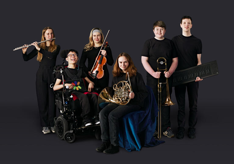 Musicians from the National Open Youth Orchestra with their instruments, in front of a dark grey background.