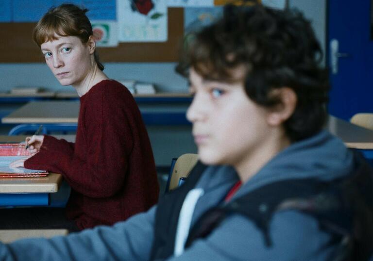 A teacher looks across the desks at a young boy student. 