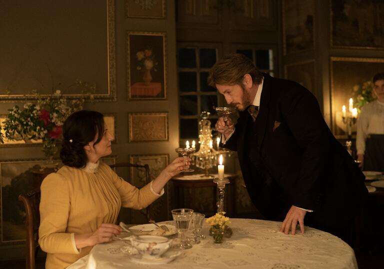 A man stands in front of a woman in a candlelit dining room, in soft evening lighting.
