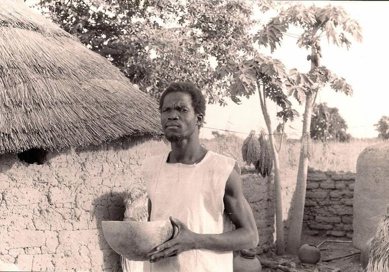 A man stands in front of a thatched roof mud hut, holding a clay pot, with a confused look on his face.
