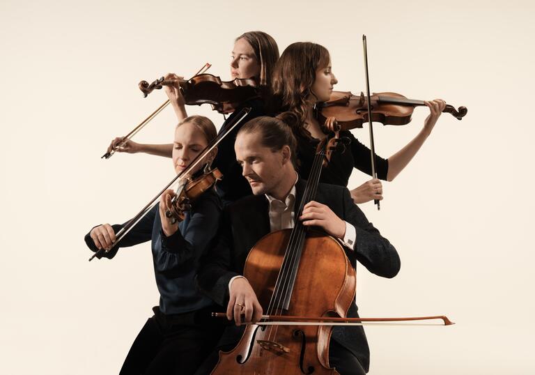 The Dudok Quartet playing their instruments. They are positioned in a close huddle, each person facing a different direction. They are against a cream coloured background.