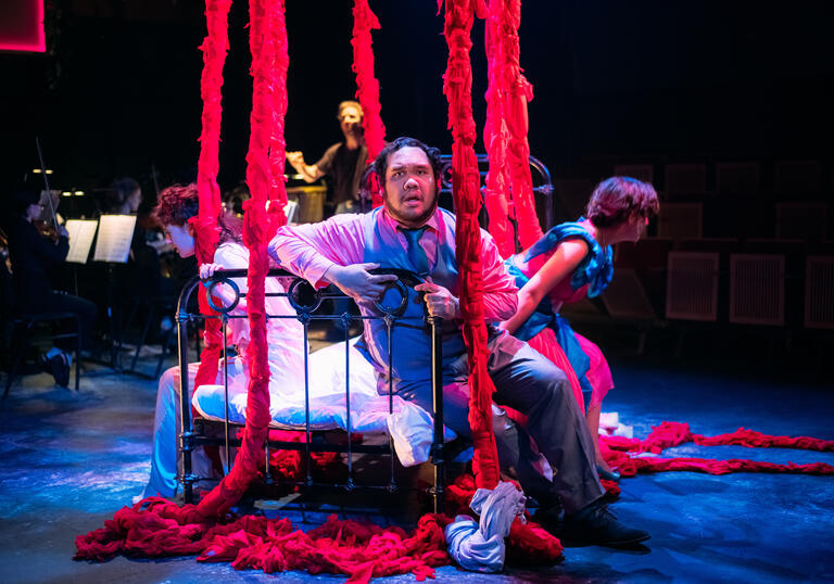 Three opera singers sat on a bed with red rope-like hanging down from ceiling. Small ensemble performing in background