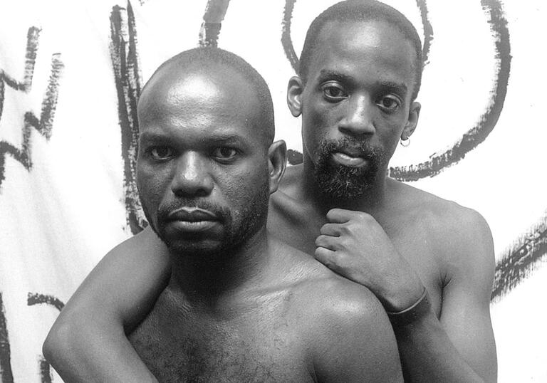 Two topless men embrace each other.