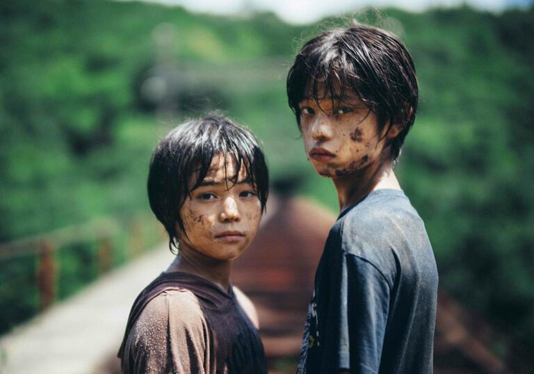 Two young boys covered in mud stand on train tracks in a rural setting, looking towards the camera.