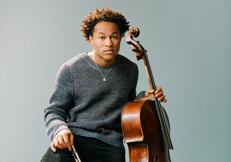 Sheku Kanneh-Mason sitting on a wooden stool with his cello, in front of a pale blueish gray background