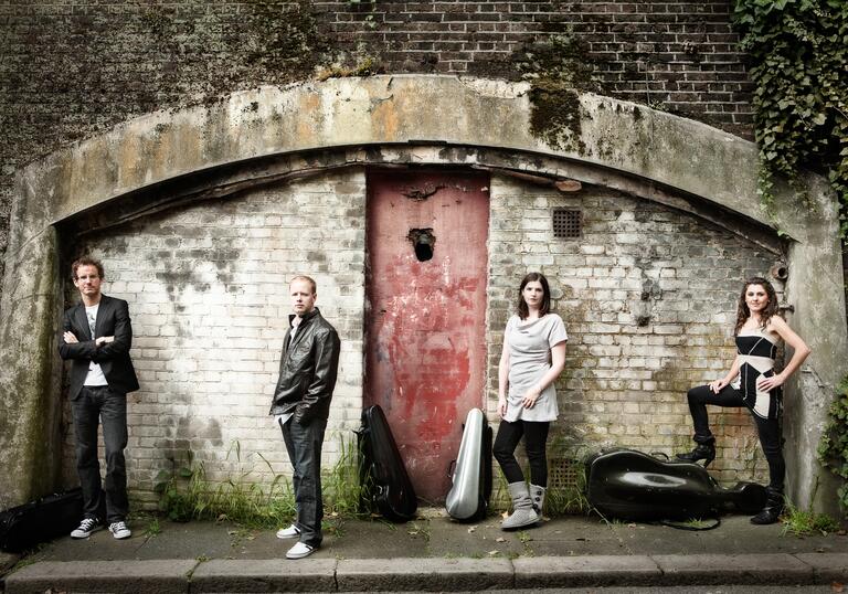 The Carducci Quartet standing in a line in front of an old, dilapidated building, their instruments in cases on the floor beside them.