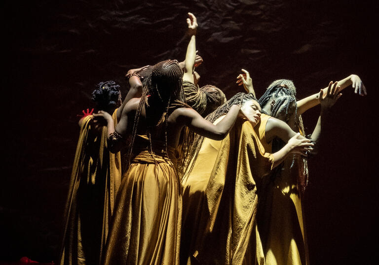 Members of the cast of The Golden Stool in long golden dresses, standing in a close huddle, some with their arms outstretched