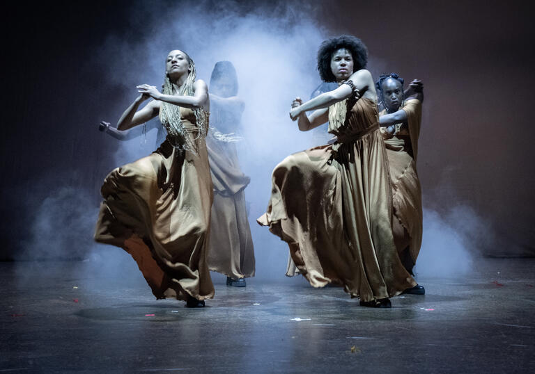 Members of the cast of The Golden Stool dancing in long golden dresses, a plume of smoke billowing from behind them